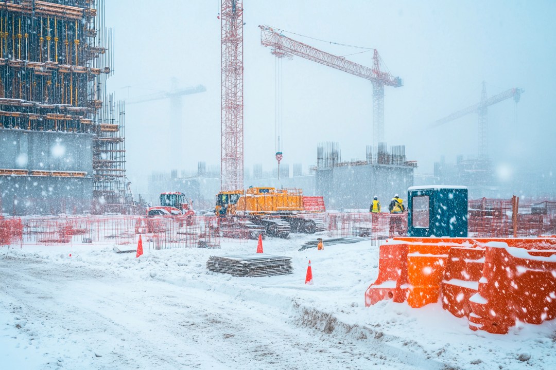 Active construction site in Canada during heavy snowfall with cranes, workers in safety gear, and snow-covered equipment showing cold-weather project conditions